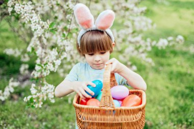 Parlak Plastik Yumurta Taşıyan Çocuk ile Paskalya Yumurtası Avı. Gülümseyen çocuk tavşan kulağı takıyor ve çiçek açan bahar bahçesinde renkli paskalya yumurtalarıyla dolu hasır bir sepet tutuyor.