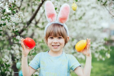 Parlak Plastik Yumurta Taşıyan Çocuk ile Paskalya Yumurtası Avı. Gülümseyen çocuk tavşan kulağı takıyor ve çiçek açan bahar bahçesinde renkli paskalya yumurtalarıyla dolu hasır bir sepet tutuyor.