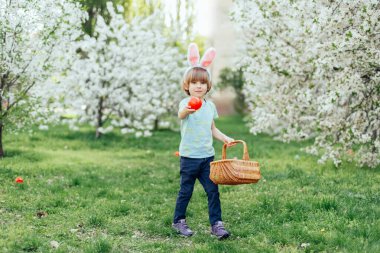 Parlak Plastik Yumurta Taşıyan Çocuk ile Paskalya Yumurtası Avı. Gülümseyen çocuk tavşan kulağı takıyor ve çiçek açan bahar bahçesinde renkli paskalya yumurtalarıyla dolu hasır bir sepet tutuyor.
