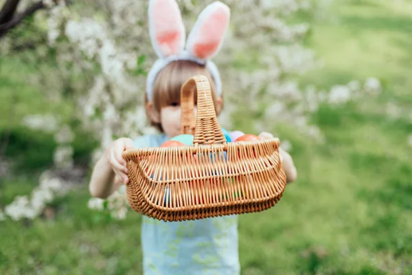 Parlak Plastik Yumurta Taşıyan Çocuk ile Paskalya Yumurtası Avı. Gülümseyen çocuk tavşan kulağı takıyor ve çiçek açan bahar bahçesinde renkli paskalya yumurtalarıyla dolu hasır bir sepet tutuyor.