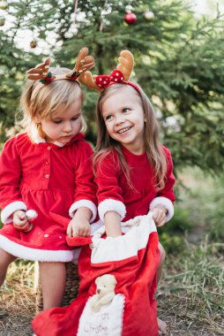Kırmızı Noel elbiseleri ve şenlikli ren geyiği boynuzları takan iki genç kız dışarıda, dekore edilmiş bir ağacın yanında Noel hediyesi çuvalı keşfediyorlar..