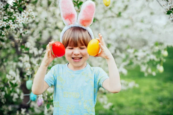 Parlak Plastik Yumurta Taşıyan Çocuk ile Paskalya Yumurtası Avı. Gülümseyen çocuk çiçek açan bahar bahçesinde tavşan kulağı takıyor.