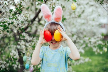 Parlak Plastik Yumurta Taşıyan Çocuk ile Paskalya Yumurtası Avı. Gülümseyen çocuk çiçek açan bahar bahçesinde tavşan kulağı takıyor.