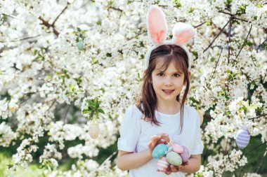 Paskalya yumurtası avı. Bahçede yumurta toplamak için koşan tavşan kulaklı bir kız çocuğu. Paskalya geleneği. Renkli yumurtalarla dolu sepeti olan bebek.