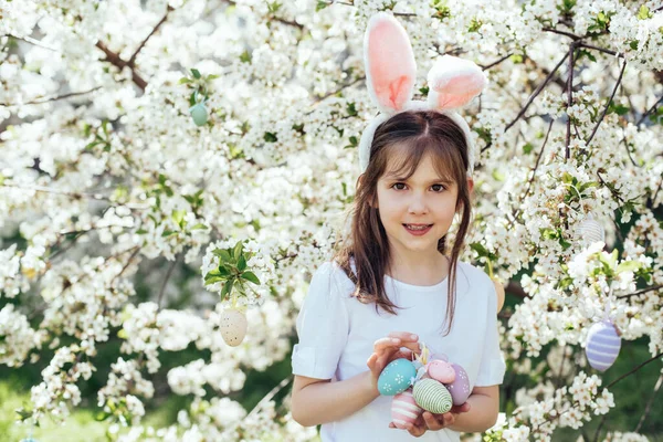 Paskalya yumurtası avı. Bahçede yumurta toplamak için koşan tavşan kulaklı bir kız çocuğu. Paskalya geleneği. Renkli yumurtalarla dolu sepeti olan bebek.