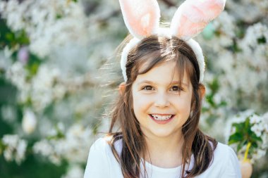 Paskalya yumurtası avı. Bahçede yumurta toplamak için koşan tavşan kulaklı bir kız çocuğu. Paskalya geleneği. Renkli yumurtalarla dolu sepeti olan bebek.