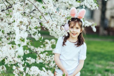 Paskalya yumurtası avı. Bahçede yumurta toplamak için koşan tavşan kulaklı bir kız çocuğu. Paskalya geleneği. Renkli yumurtalarla dolu sepeti olan bebek.