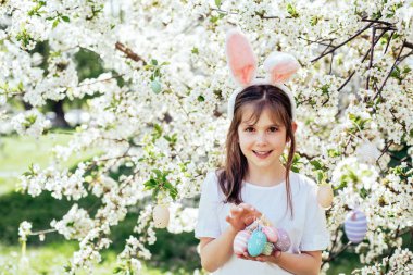 Paskalya yumurtası avı. Bahçede yumurta toplamak için koşan tavşan kulaklı bir kız çocuğu. Paskalya geleneği. Renkli yumurtalarla dolu sepeti olan bebek.