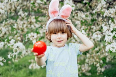Parlak Plastik Yumurta Taşıyan Çocuk ile Paskalya Yumurtası Avı. Gülümseyen çocuk tavşan kulağı takıyor ve çiçek açan bahar bahçesinde renkli paskalya yumurtalarıyla dolu hasır bir sepet tutuyor.