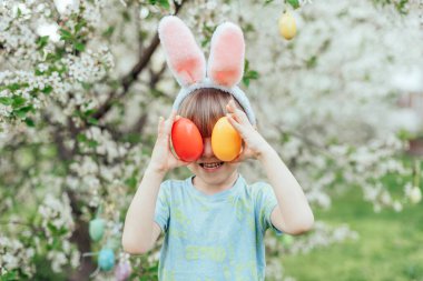Parlak Plastik Yumurta Taşıyan Çocuk ile Paskalya Yumurtası Avı. Gülümseyen çocuk tavşan kulağı takıyor ve çiçek açan bahar bahçesinde renkli paskalya yumurtalarıyla dolu hasır bir sepet tutuyor.