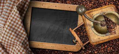 Metal ve ahşaptan yapılmış eski el yapımı kahve öğütücüsü masada kavrulmuş kahve çekirdekleri boş bir karatahta ve kareli bir masa örtüsü..