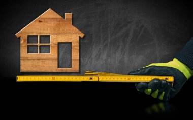 İş eldivenli bir elin yakın çekimi. Sarı ahşap bir cetvel tutuyor. Tahtasında tahta bir model ev var..