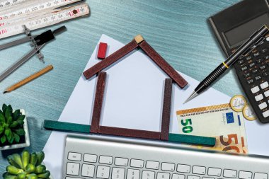 Masanın üzerinde oyuncak bloklardan yapılmış küçük tahta bir ev, Euro para birimi, hesap makinesi, katlama cetveli, kalem ve bir çizim pusulası var. Gayrimenkul yatırımları kavramı.  