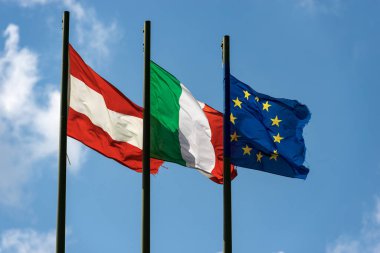 Avusturya, İtalya ve Avrupa Birliği bayrakları bayrak direğiyle birlikte açık mavi gökyüzünde bulutlar ve kopyalama alanlarıyla birlikte rüzgarda dalgalanır..