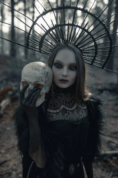 Young woman in image of witch stands in black dress and crown on her head with human skull in her hand in dark forest.