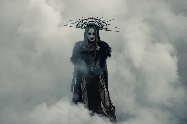 Young woman in image of witch stands in black dress and crown on her head against smoke background.