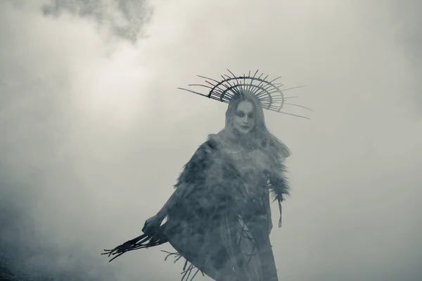 Young woman in image of witch stands in black dress and crown on her head against smoke background.