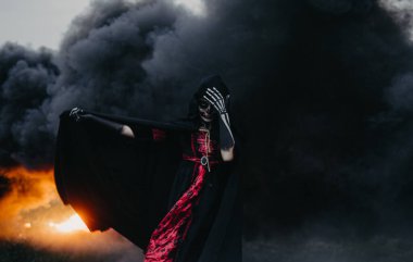 Genç bir kadın, Cadılar Bayramı ölüm kostümü içinde vücudunda boyalı bir iskeletle ateş, alev ve siyah dumanla sahada duruyor. Cadılar Bayramı konsepti Şeker kafatası makyajı.