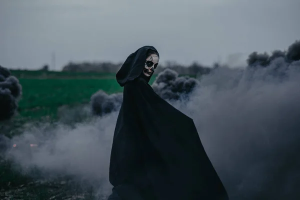 若い女性は黒煙を背景に死のハロウィーンの衣装のフィールドに立っています ハロウィンのコンセプト 砂糖の頭蓋骨のメイク — ストック写真