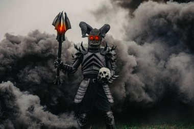 Mutant savaşçı parlayan gözleriyle ayakta duruyor ve elinde bir topuzla bir insan kafatasını siyah bir dumana karşı tutuyor..