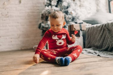 Çocuk Noel süsüsüyle yerde oturuyor ve elinde lolipopla yatağın yanında Noel ağacı tutuyor..