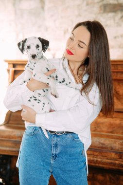 Genç kadın piyano yanındaki odada dalmaçyalı köpek yavrusuyla duruyor..