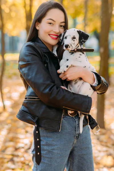 Young woman holds puppy of dalmatian dog in her hands while walking in autumn park.