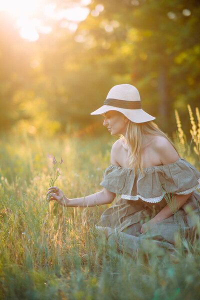 Young happy woman sits and enjoys in dress and hat in meadow at sunset. Backlit.