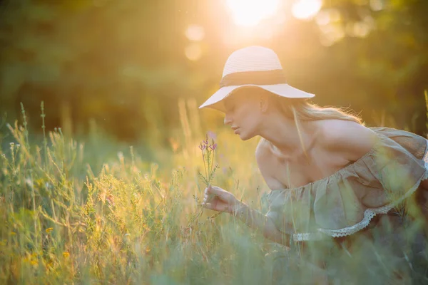 若い幸せな女性が座って 楽しんで 日没時にドレスや牧草地で帽子で野の花を滑ります バックライト付き — ストック写真