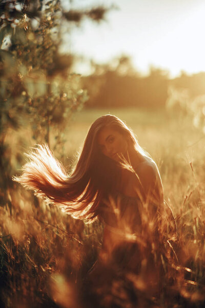 Young happy woman in dress sits and enjoys in meadow at sunset. Backlit.