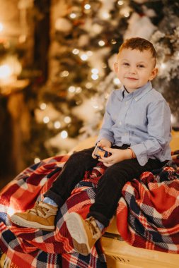 Küçük bir çocuk eski bir arabanın kaputunda oturuyor. Arka planda bir Noel ağacı ve süslemeleri var..