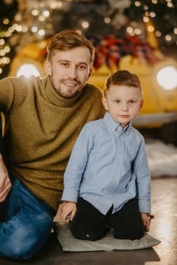 Mutlu baba ve oğlu yerde oturup arka planda parlayan retro araba farlarına ve ışıklı Noel süslemelerine karşı oynuyorlar..