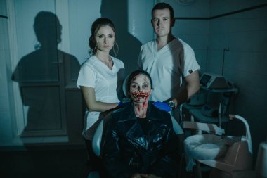 Çirkin, kanlı mutant görünümlü bir kadın dişçi ve asistanının yanındaki modern diş kliniğinde sandalyede oturuyor. KostümComment.