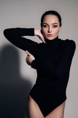 Genç bir kadın, gri arka planda gölgesinin yanında siyah bir elbise içinde poz veriyor..