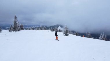 Snowboardcu, kozalaklı ağaçların arasında karlı yamaçlara çıkar ve bulutların ve dağların arkasına kar yağar. İHA ateşi.