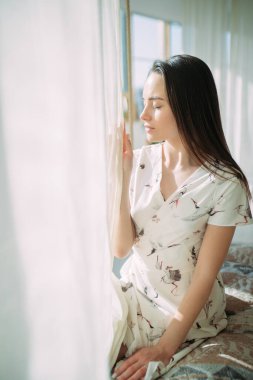 Genç bir kadın sabahlıkla yatakta oturur ve pencerenin yanında meditasyon yapar..