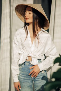 Genç bir kadın rahat ve gözleri kapalı duruyor kot pantolon, beyaz gömlek ve Vietnam şapkası içinde..