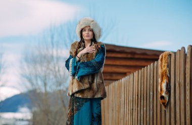 Kürk şapka, yelek ve elbise giymiş genç bir kadın üzerinde tilki postu asılı ahşap çitin yanında duruyor..