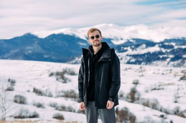 Genç sakallı adam ceketli ve güneş gözlüklü, karla kaplı Karpatlar 'ın güzel manzarasına karşı duruyor..