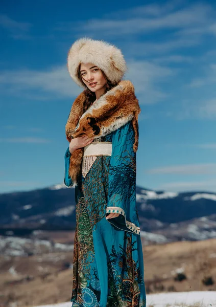 Young woman stands dressed in fur hat, dress and collar fox against beautiful landscape of snowbound Carpathians mountains and sky.