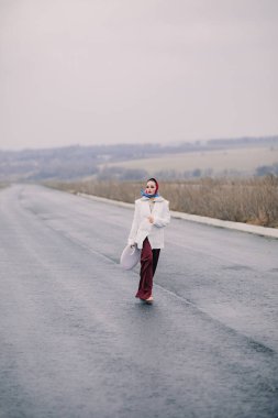 Genç zarif kadın otobanda yürüyor. Kırsal kesimde pantolon, ceket ve şal giyiyor..