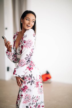 Genç bir kadın yazın şık bir çiçek desenli elbise giyer, elinde akıllı bir telefon ve neşeli bir gülümsemeyle durur..