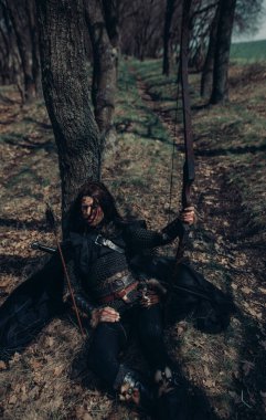 Woman in chain mail in image of medieval warrior sits and rests near tree among forest. clipart