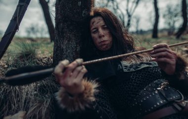 Zincirli kadın ortaçağ savaşçısı görüntüsünde yatıyor, dinleniyor ve okunu ormanın yakınındaki ağaçların arasında inceliyor..