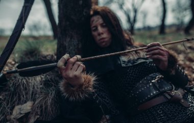 Ortaçağ savaşçısı görüntüsüne bürünmüş zincirli bir kadın ormanın yakınındaki ağaç yakınlarında yatıyor, dinleniyor ve okunu inceliyor. Eline odaklan..