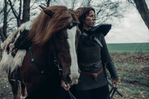 中世纪战士形象中戴着铁链的女人站在马旁边 握在森林里的缰绳上 — 图库照片
