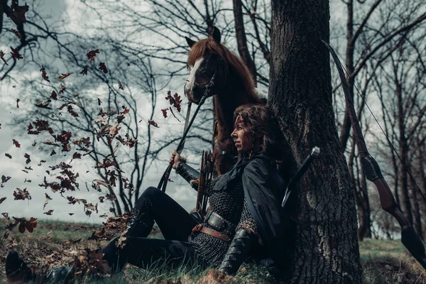Woman in chain mail in image of medieval warrior sits and rests near tree among forest and holds horse by the reins.