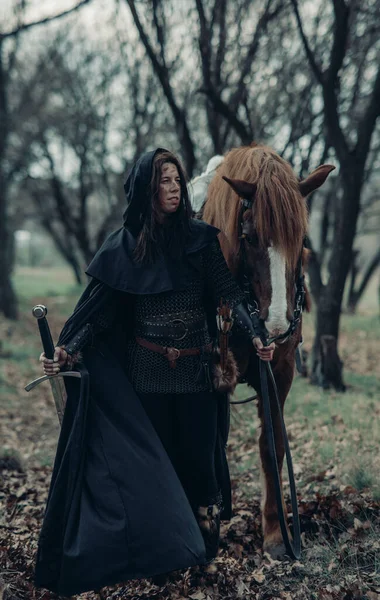 Woman in chain mail in image of medieval warrior leads her horse by the reins among forest with sword in her hand.