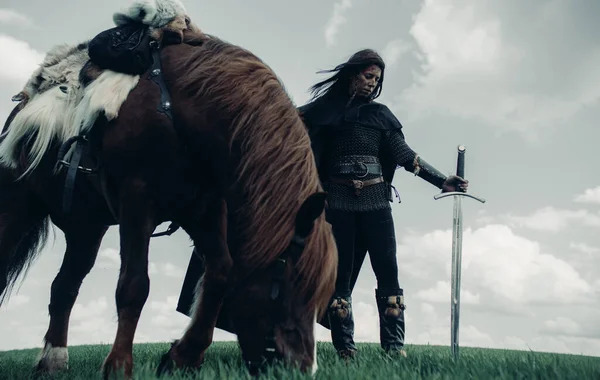 Woman in chain mail in image of medieval warrior stands with sword in her hand near horse in field.