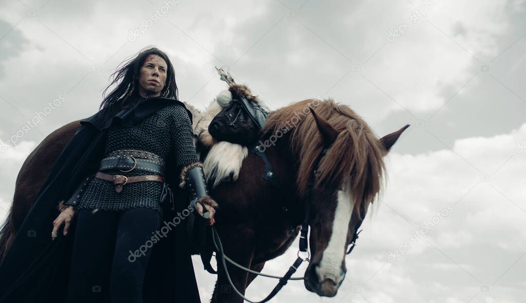 Woman in chain mail in image of medieval warrior stands near her horse against background of sky and clouds.